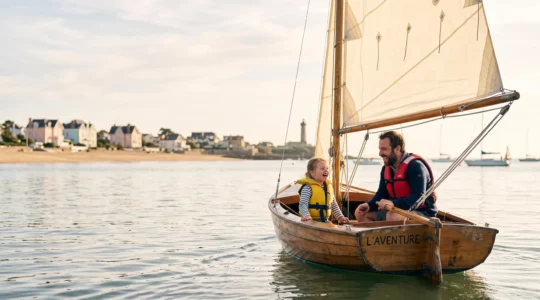 Un parent et son enfant naviguent ensemble sur un petit dériveur dans une baie calme de Loire-Atlantique, avec la côte en arrière-plan